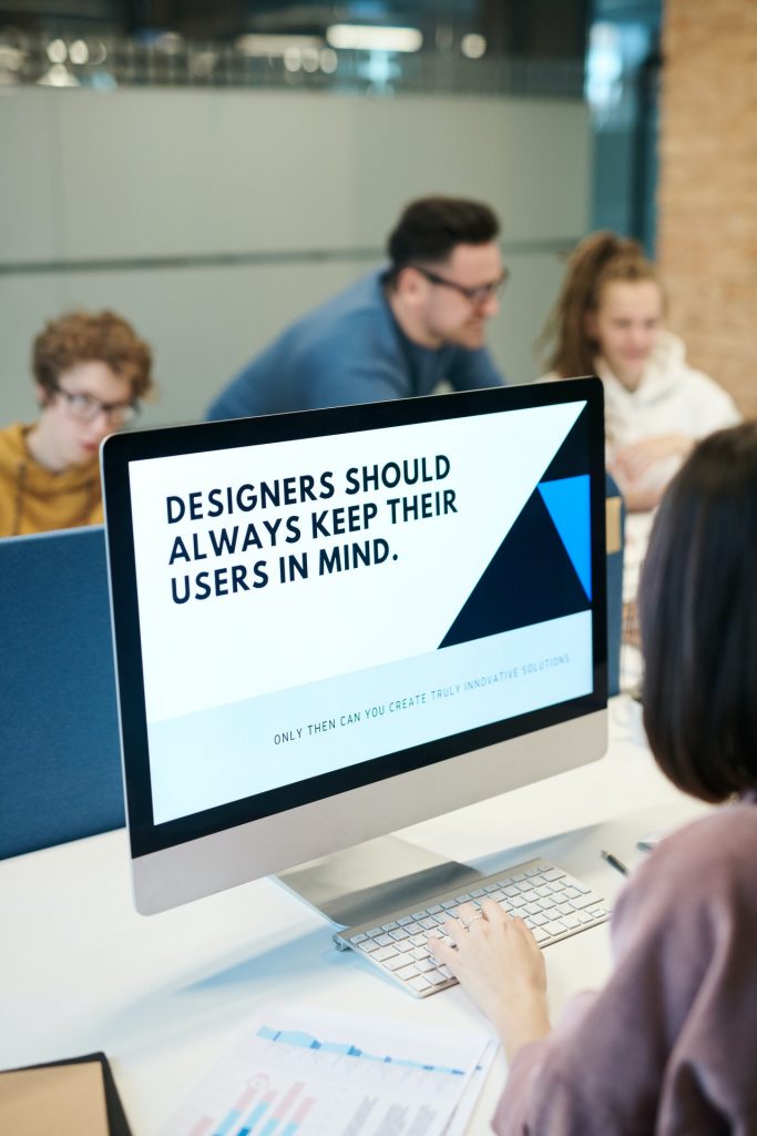 Room of people with one person looking at a laptop. The laptop screen reads "Designers should always keep their users in mind."